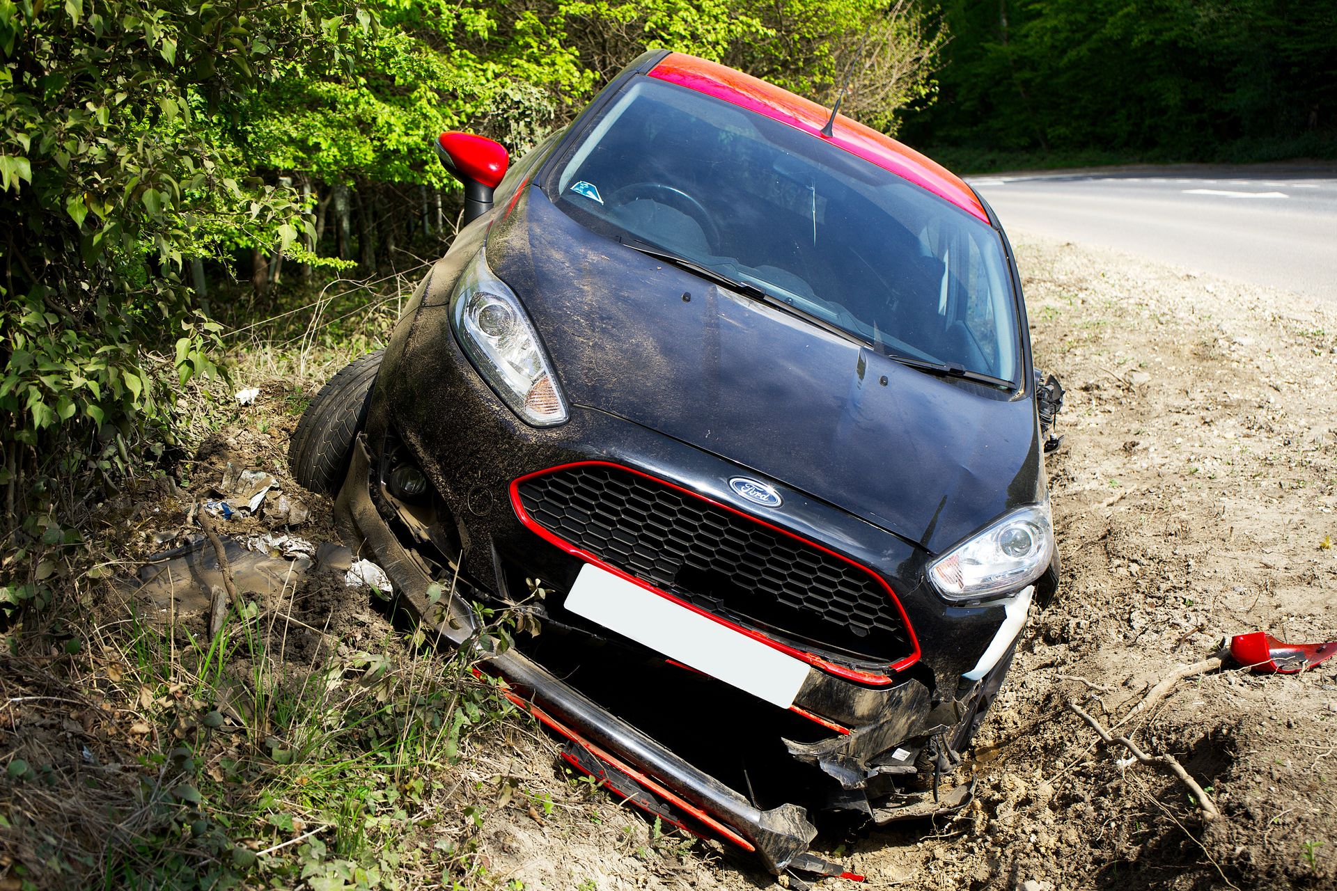 A black car, seemingly a Ford, crashed and damaged in a roadside ditch with surrounding greenery.